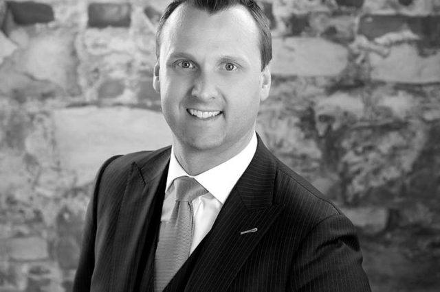 portrait fo man with short hair in suit in front of brick wall